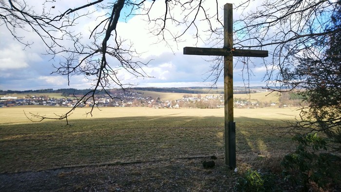 Ein hölzernes Kreuz steht auf einem Hügel mit Blick auf ein Dorf.