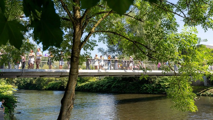 Menschen gehen an einem sonnigen Tag über eine malerische Brücke über einen ruhigen, von Bäumen gesäumten Fluss.