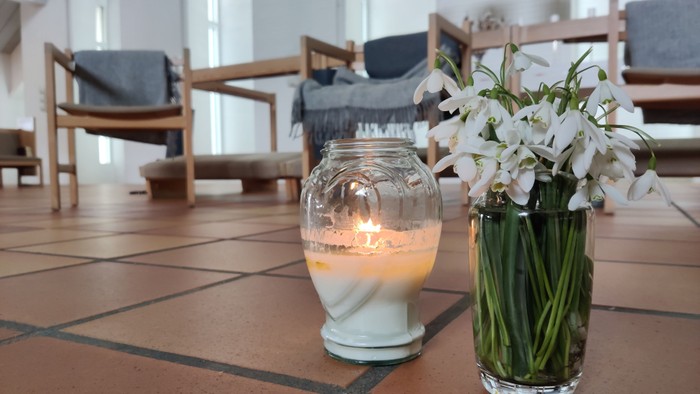 Et tændt stearinlys og en vase med hvide blomster står på et flisebelagt bord i et rum med træstole.