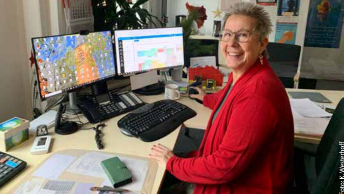 Eine lächelnde Frau in einem roten Pullover arbeitet an einem Schreibtisch mit mehreren Monitoren und verschiedenen Büroartikeln.