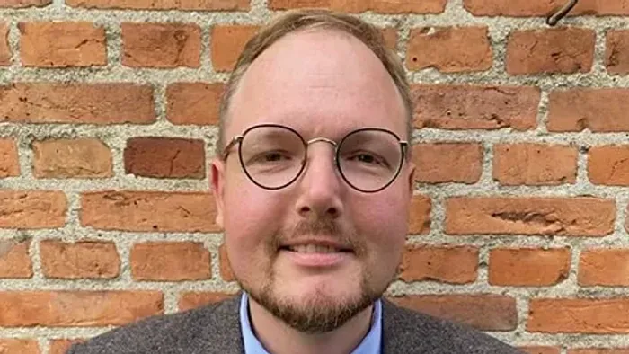 En mand med briller, grå blazer, blå skjorte og mønstret butterfly står op ad en murstensvæg.