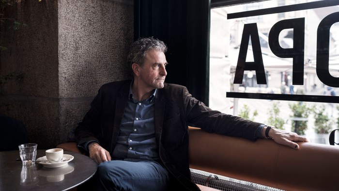 En mand sidder eftertænksomt ved et vindue på en café og nipper til sin kaffe med en afslappet holdning.