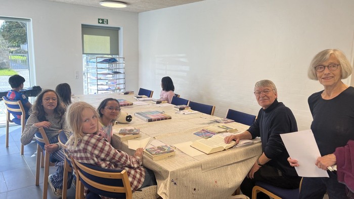 En gruppe af voksne sidder ved et bord i et rum med blå stoler og et hvidt væg.