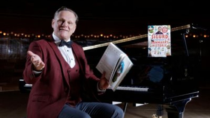 En mand i rødt jakkesæt sidder ved et piano og holder en bog.