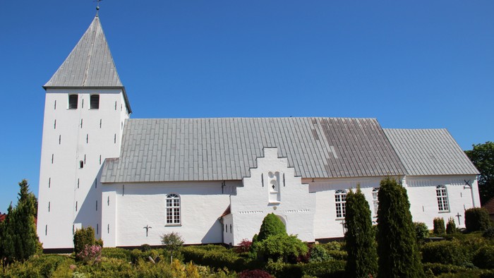 Nørre Løgum kirke, set fra syd med blå himmel som baggrund. Kirken er hvidkalket, har et tårn og der er bly på taget. Forrest ses grønnehække og buske på kirkegården