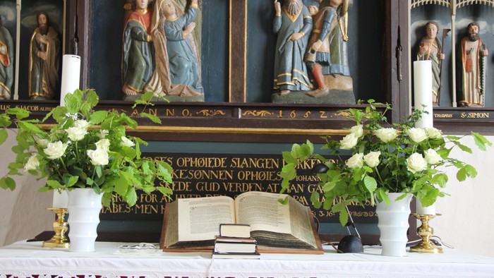 Nærbillede af Nr. Løgum kirkes alterbord med den hvide alterdug. På begge sider af bordet ses de hvide alterlys i messingstager og foran dem store vaser med bøgegrene og hvide roser. I midten ses en stor opslået Bibel med et par små bøger foran. Det neder