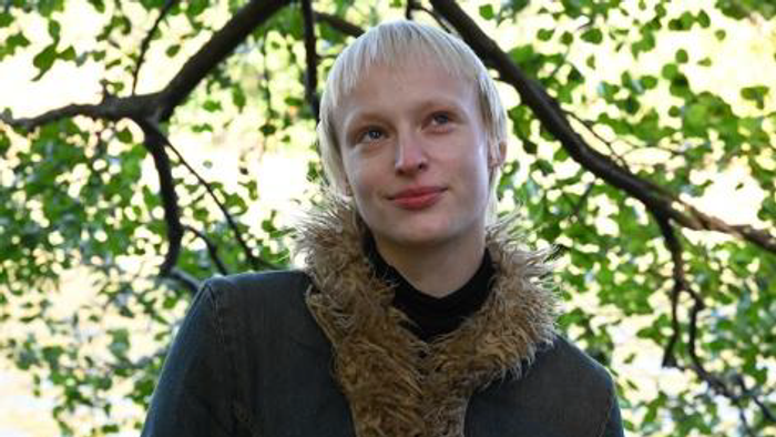 Person mit blondem Haar, die einen dunklen Mantel und einen pelzgefütterten Schal vor einem grünen Baumhintergrund trägt.