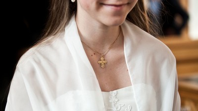 En person i en hvid blondebluse bærer et guldkors-halskæde og smiler blidt.