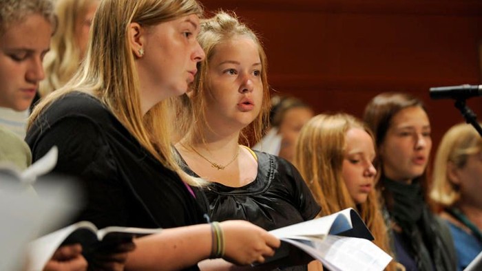 Eine Gruppe junger Frauen, die gemeinsam in einem Chor singen und sich auf ihre Notenblätter konzentrieren.