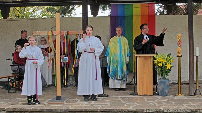 Ein Priester predigt von einem Podium, umgeben von Kirchenmitgliedern und Symbolen.