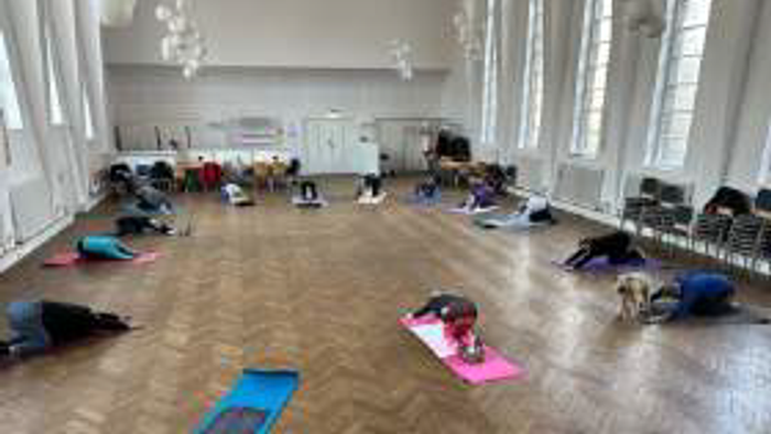 Gruppe, die Yoga auf Matten in einem geräumigen Innenraum mit Holzboden und Stühlen drumherum praktiziert.