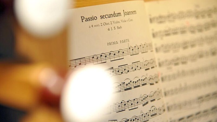 Nahaufnahme klassischer Notenblätter mit dem Titel *„Passio secundum Joannem“* von J.S. Bach, unscharfe dekorative Lichter im Hintergrund.