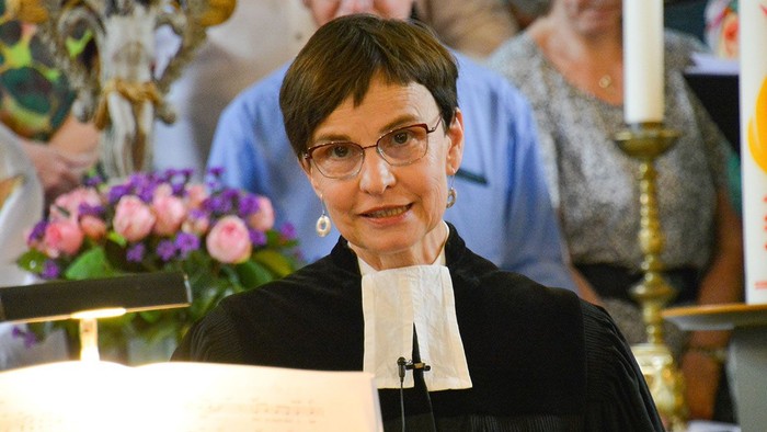 Pfarrerin in schwarzer Amtstracht mit weißem Beffchen steht am Lesepult in einer Kirche und spricht, im Hintergrund sind unscharf weitere Menschen sowie Kerzen und Blumenschmuck zu sehen.