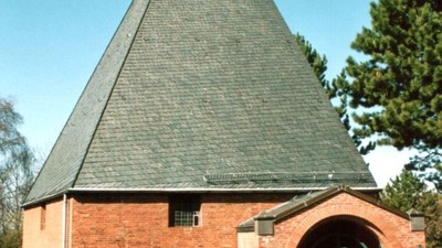 Idyllische Backsteinkapelle mit steilem dreieckigem Dach und markantem Kreuz an der Spitze.