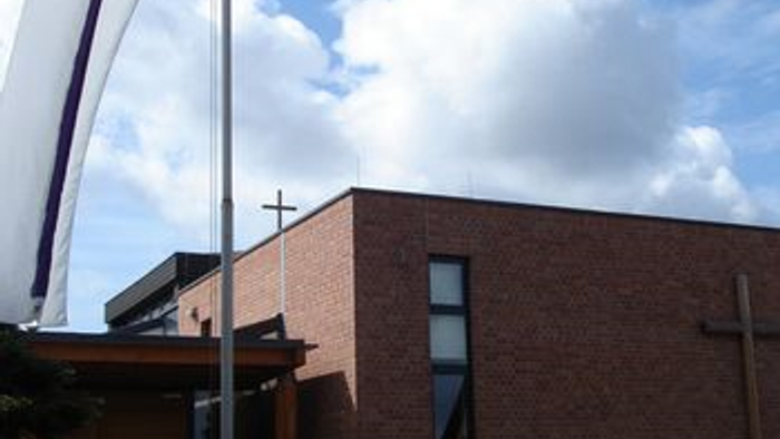 Moderne Backsteinkirche mit einem Fahnenmast, der ein Kreuzbanner zeigt, und einem gepflegten Gartenweg.