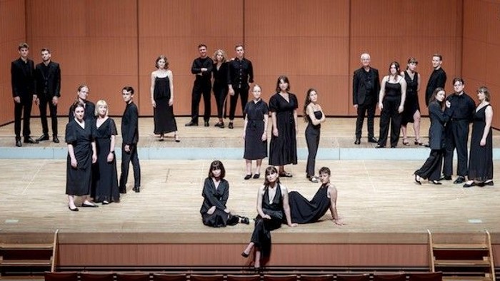 En gruppe optrædende i formelt tøj poser dramatisk på en scene, hvor de blander stående og siddende stillinger.