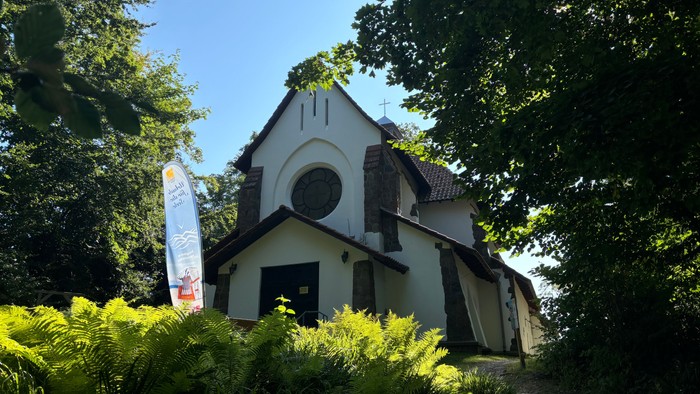 Kleine weiße Kirche mit Rundbogenfenster und Kreuz, umgeben von Bäumen und Büschen