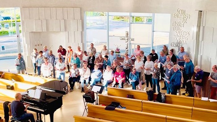 En gruppe synger og spiller instrumenter i en rummelig, veloplyst auditorium med en pianist forrest.