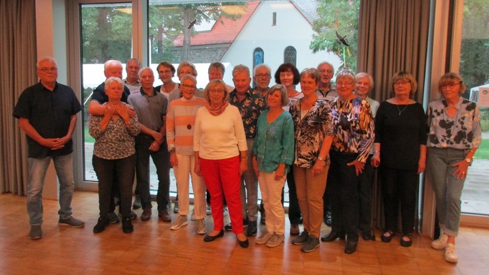 Eine Gruppe älterer Menschen posiert für ein Gruppenfoto in einem Raum mit großen Fenstern.