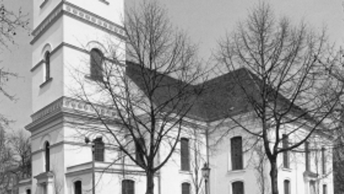 Historische Kirche mit hohem Glockenturm und kahlen Bäumen in einer schwarz-weißen Stadtkulisse.