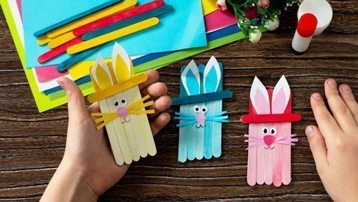Drei Bastelhasen aus Eisstielen, gehalten von Händen, mit bunten Bastelmaterialien im Hintergrund.