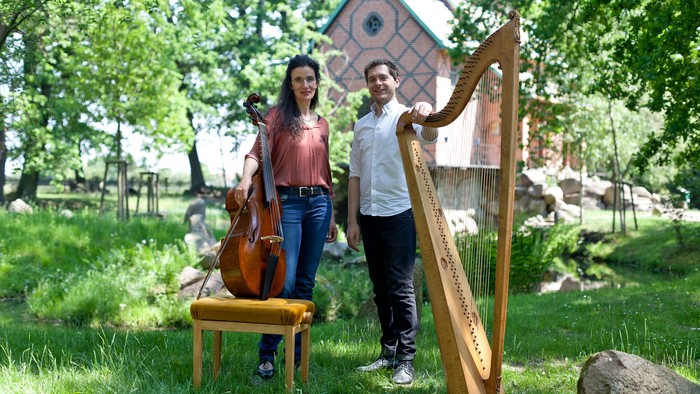 Zwei Musiker posieren im Freien mit einem Cello und einer Harfe in einer üppigen, grünen Parklandschaft.
