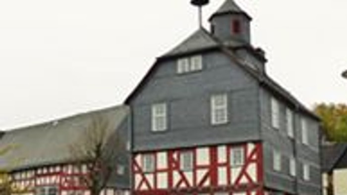 Großer Scheunenbau mit rot-weißer Fachwerkkonstruktion, grauer Dachbedeckung und einer Wetterfahne auf dem Dachfirst.