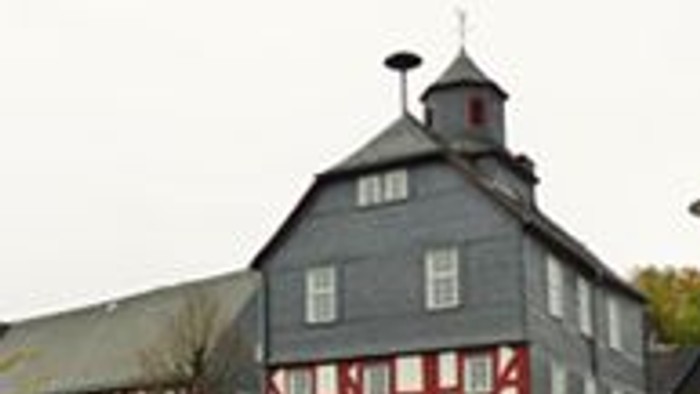 Großer Scheunenbau mit rot-weißer Fachwerkkonstruktion, grauer Dachbedeckung und einer Wetterfahne auf dem Dachfirst.