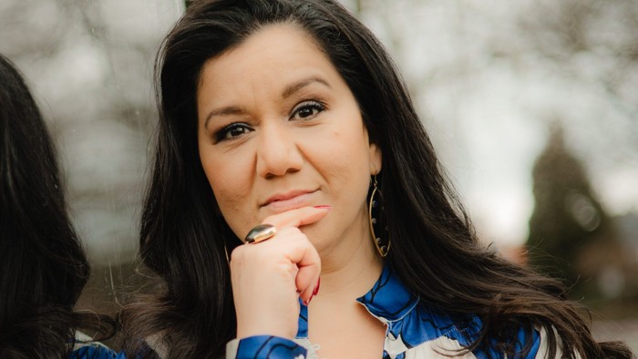 En kvinde i en blå og hvidmønstret bluse hviler eftertænksomt hagen i hånden udendørs.