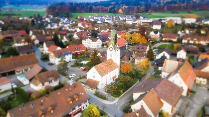 Dorf mit Kirche und umliegenden Häusern, umgeben von Bäumen und Feldern