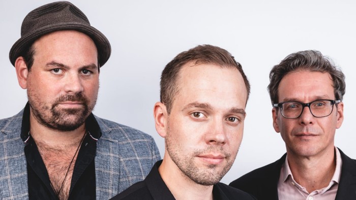 Drei Männer posieren für ein Werbefoto des LoveBird Festivals neben dem bunten Logo und dem Veranstaltungsnamen.