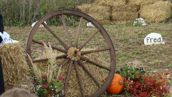 Landlig høstscene med en vognhjul, høballer, græskar, blomster og en gravsten med navnet