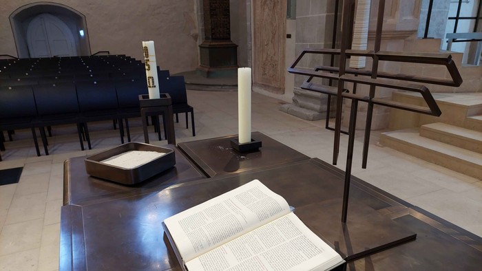 Ein offenes Buch liegt auf einem hölzernen Lesepult in einer ruhigen, schwach beleuchteten Kapelle mit religiöser Ausstattung.