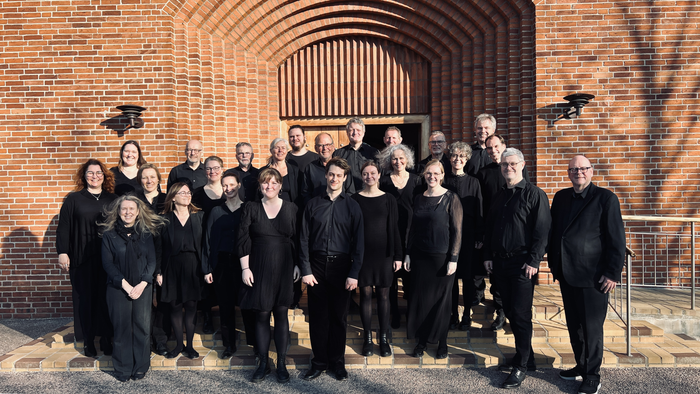 En gruppe musikere i formelt sort tøj poser sammen foran indgangen til en murstensbygning.