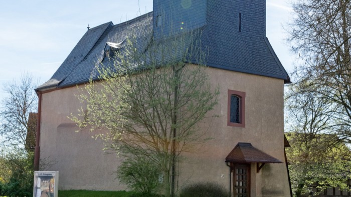 Kirche mit hohem Turm und Kreuz, umgeben von Bäumen und Gras.