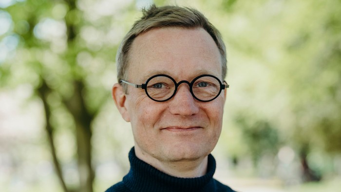 En person iført runde briller og en mørk halsturtleneck-sweater udendørs i dagslys.