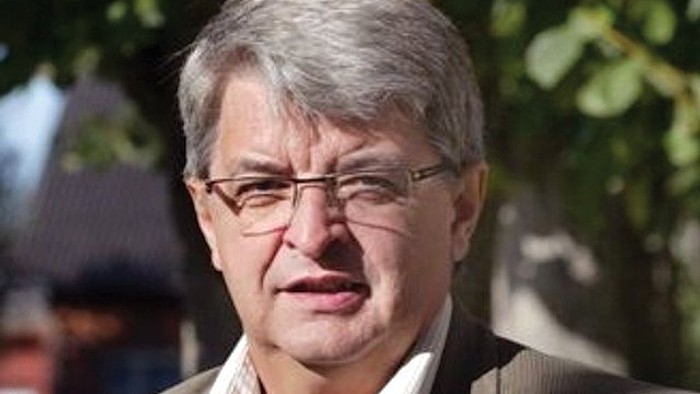 En mand med briller og kort gråt hår, iført en jakke og en lys skjorte udendørs.