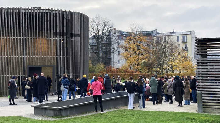 Menschen versammeln sich vor modernen architektonischen Gebäuden mit Holzverkleidung an einem bewölkten Tag.