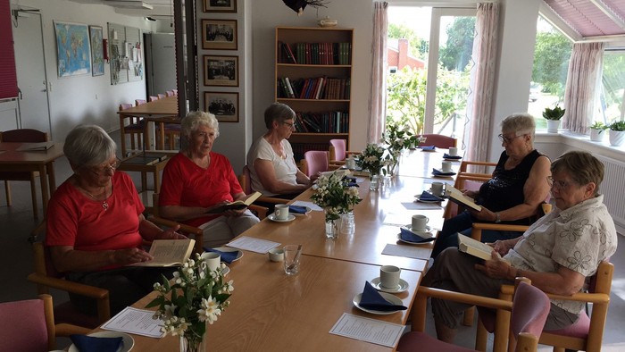 En gruppe pensionister sidder ved et langt bord og spiser morgenmad i et lys og behageligt rum med blomster og bøger.