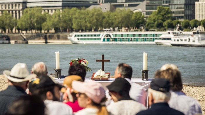 Eine Gruppe von Menschen nimmt an einer Andacht am Flussufer teil, mit Kreuz und Kerzen, im Hintergrund Boote und Gebäude.