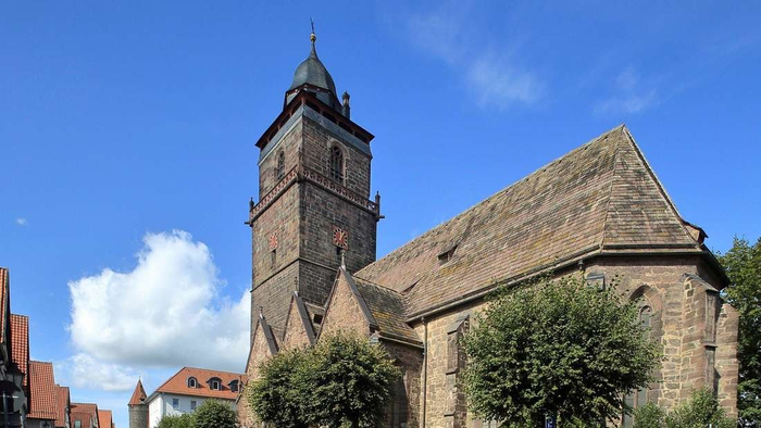 Kirche mit hohem Turm und steilem Dach, umgeben von Bäumen und anderen Gebäuden unter blauem Himmel.