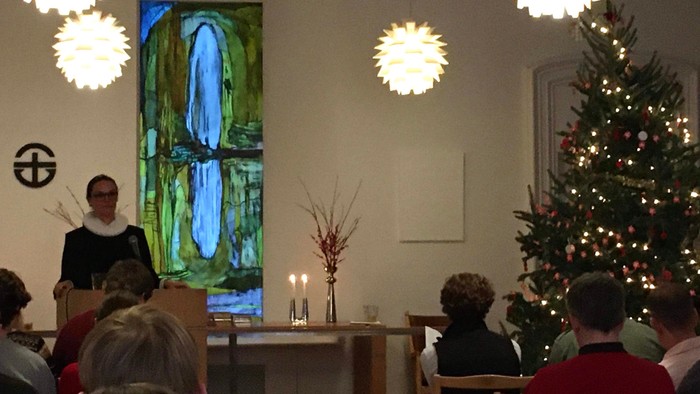 En præst holder et tale foran en lille kirkekonge med lys, mens en maleri hænger på væggen.