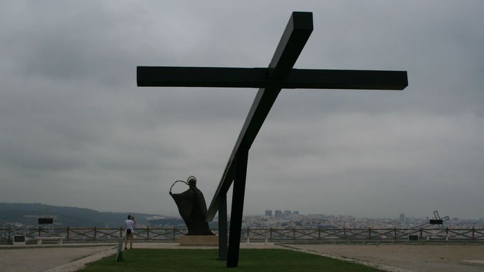 Ein großer schwarzer Kreuzmonument steht auf einem grasbewachsenen Hügel mit Stadtblick.