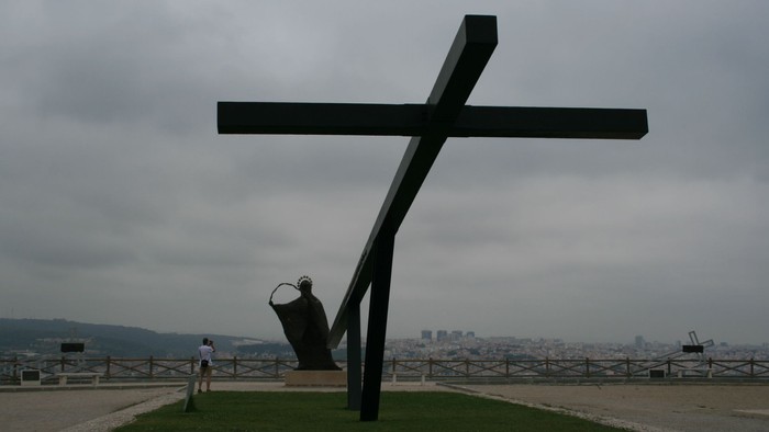 Ein großer schwarzer Kreuzmonument steht auf einem grasbewachsenen Hügel mit Stadtblick.