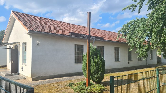 Kleines weißes Gebäude mit rotem Dach und Schornstein, umgeben von einem Zaun und Grünfläche