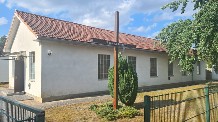 Kleines weißes Gebäude mit rotem Dach und Schornstein, umgeben von einem Zaun und Grünfläche