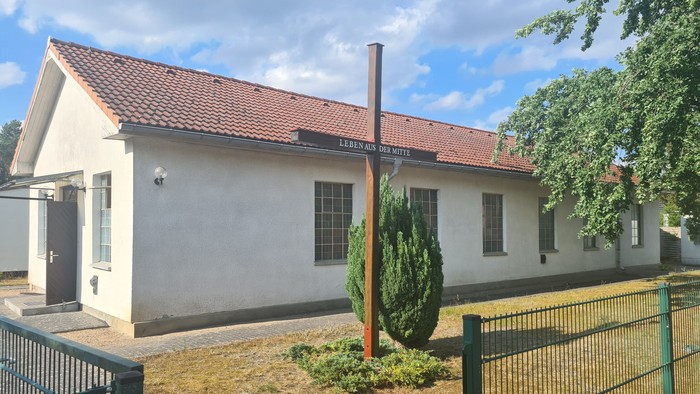 Kleines weißes Gebäude mit rotem Dach und Schornstein, umgeben von einem Zaun und Grünfläche