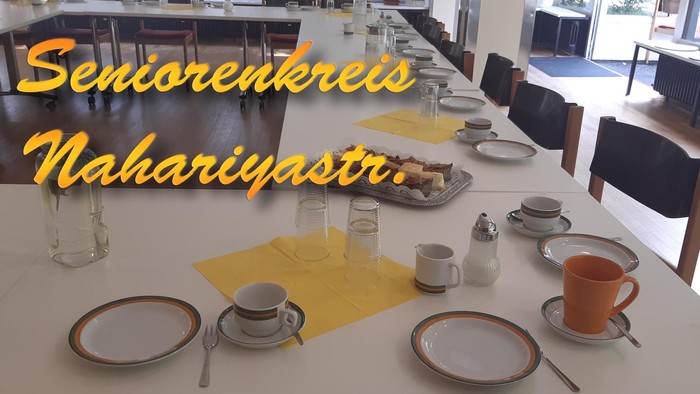 Ein ordentlich gedeckter Esstisch in einem Gemeindezentrum mit der Beschilderung „Seniorenkreis Nahariyastra“, auf dem Kaffeetassen und Besteck zu sehen sind.