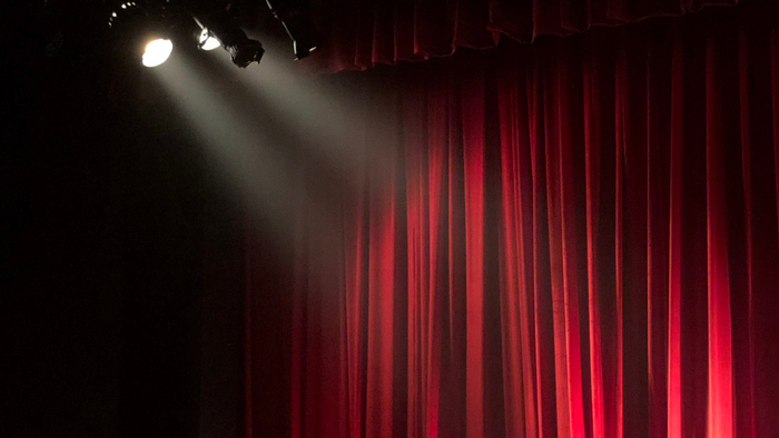 Eine Live-Bühne mit roten Vorhängen, die von einem Scheinwerfer beleuchtet wird, mit Publikum und einem Künstler.