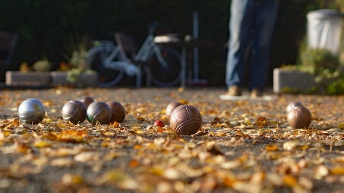 Pétanque-Kugeln verstreut auf kiesigem Boden mit Herbstblättern, eine Person spielt im Hintergrund.